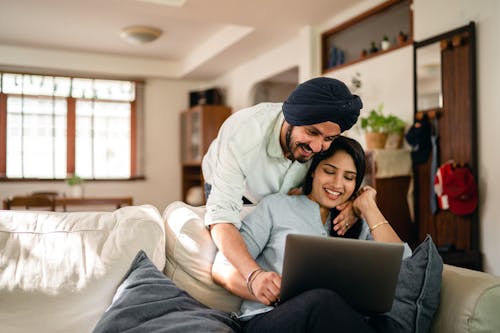 Cheerful Indian man and woman in casual clothes embracing and surfing internet on laptop while hugging in cozy living room during daytime