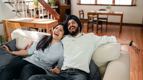 From above of optimistic young ethnic man and woman in casual outfit laughing and chilling together while embracing each other on cozy couch at home