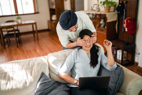 From above of young Indian woman in casual wear resting on comfortable couch with laptop in modern apartment while Indian boyfriend standing behind and closing eyes girlfriend with hands