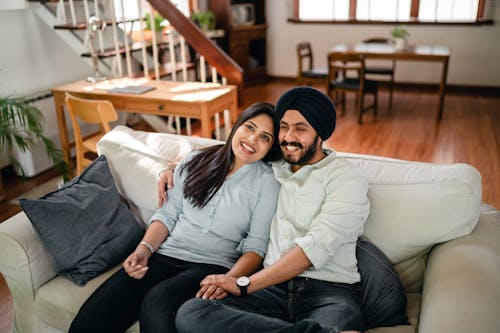 From above of smiling Indian man and woman in casual clothes resting together on comfortable couch in comfortable living room while man holding hand of girlfriend