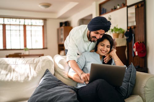 Young Indian man in turban laughing and embracing girlfriend while Indian woman sitting on sofa and browsing laptop in cozy apartment