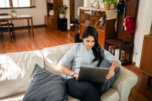 From above of young ethnic woman with long dark hair in casual clothes sitting on sofa and surfing internet on netbook while working remotely in cozy living room