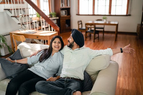 Happy adult ethnic man embracing wife and showing new housing while ethnic couple in casual clothes sitting on comfortable couch in living room