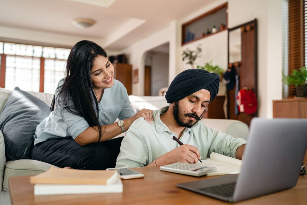 Concentrated ethnic male sitting at coffee table and writing notes near laptop and calculator and smartphone and books while smiling ethnic wife watching behind and supporting husb