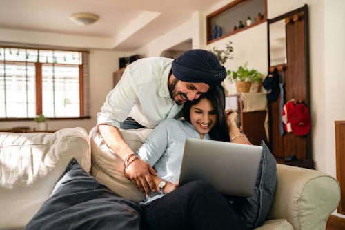 Cheerful man bending over sofa and putting hand on hand of ethnic wife sitting with laptop while looking at screen together in living room