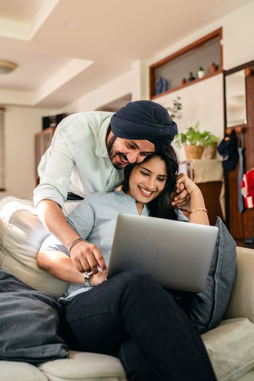 Free Positive ethnic man bending over ethnic wife using laptop on sofa and pointing on screen while resting at home together Stock Photo