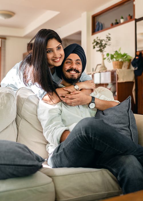 Free Positive ethnic man in casual clothes sitting on sofa hugged from back by ethnic wife while spending time together at home Stock Photo