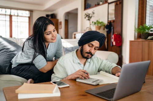 Smiling focused ethnic woman sitting on sofa at home watching working ethnic husband calculating sitting at coffee table among calculator and laptop and smartphone and books and notebook in living room