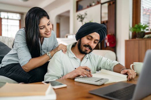 Smiling young Indian female in casual clothes supporting husband during remote work at home sitting in front of laptop with calculator notebook smartphone and cup of coffee at table