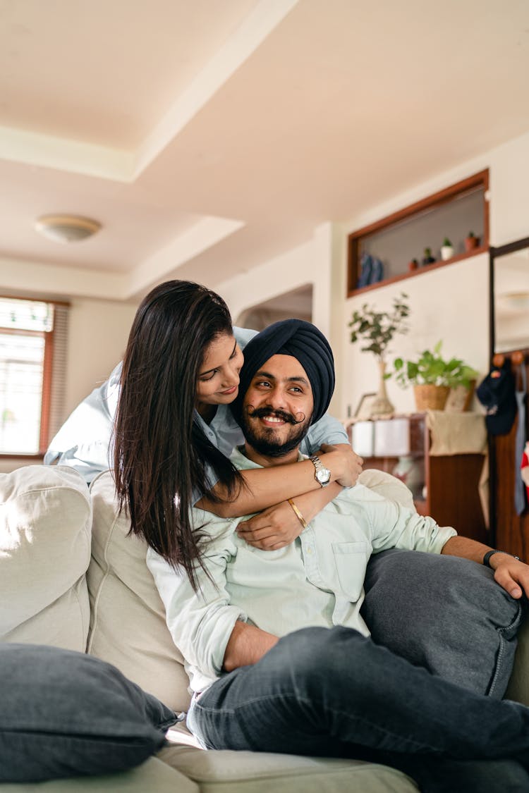 Happy Young Indian Woman Hugging Husband Sitting On Sofa At Home