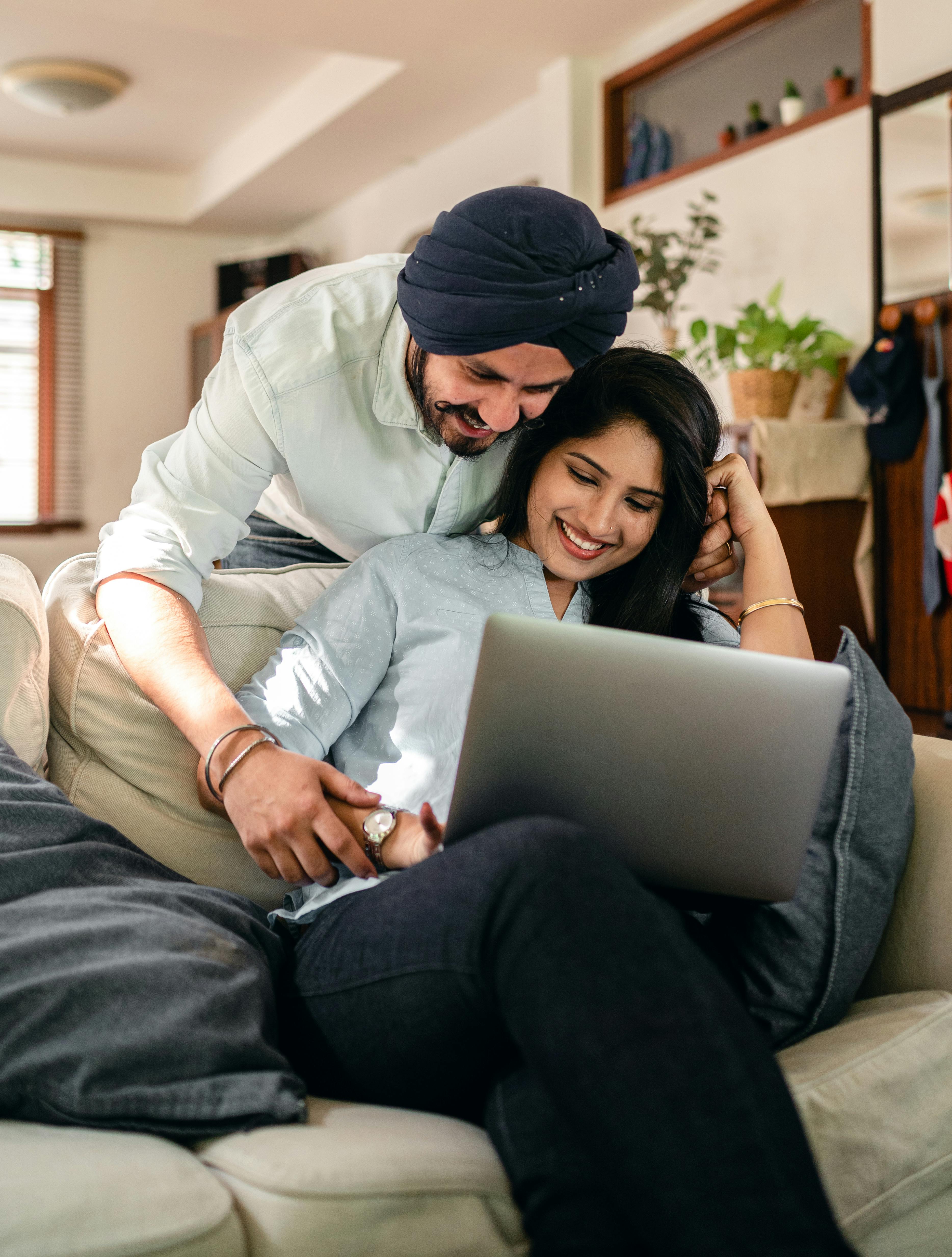 cheerful young indian man flirting with wife sitting on sofa and using laptop at home