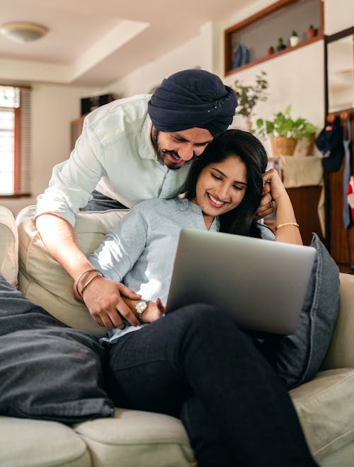 Cheerful young Indian man flirting with wife sitting on sofa and using laptop at home