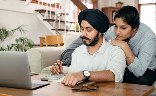 Free Focused Indian woman standing behind man in turban at table counting with calculator while writing notes in notepad Stock Photo