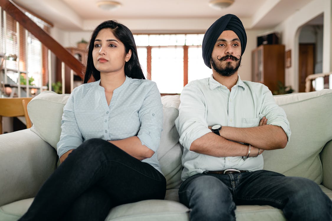 Resentful young Indian couple ignoring each other while sitting on couch together with crossed arms