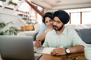 Cheerful young Indian woman cuddling and supporting serious husband working at home with laptop and counting on calculator