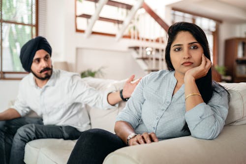 Young bearded Sikh man in casual wear and turban arguing with sad wife sitting on sofa