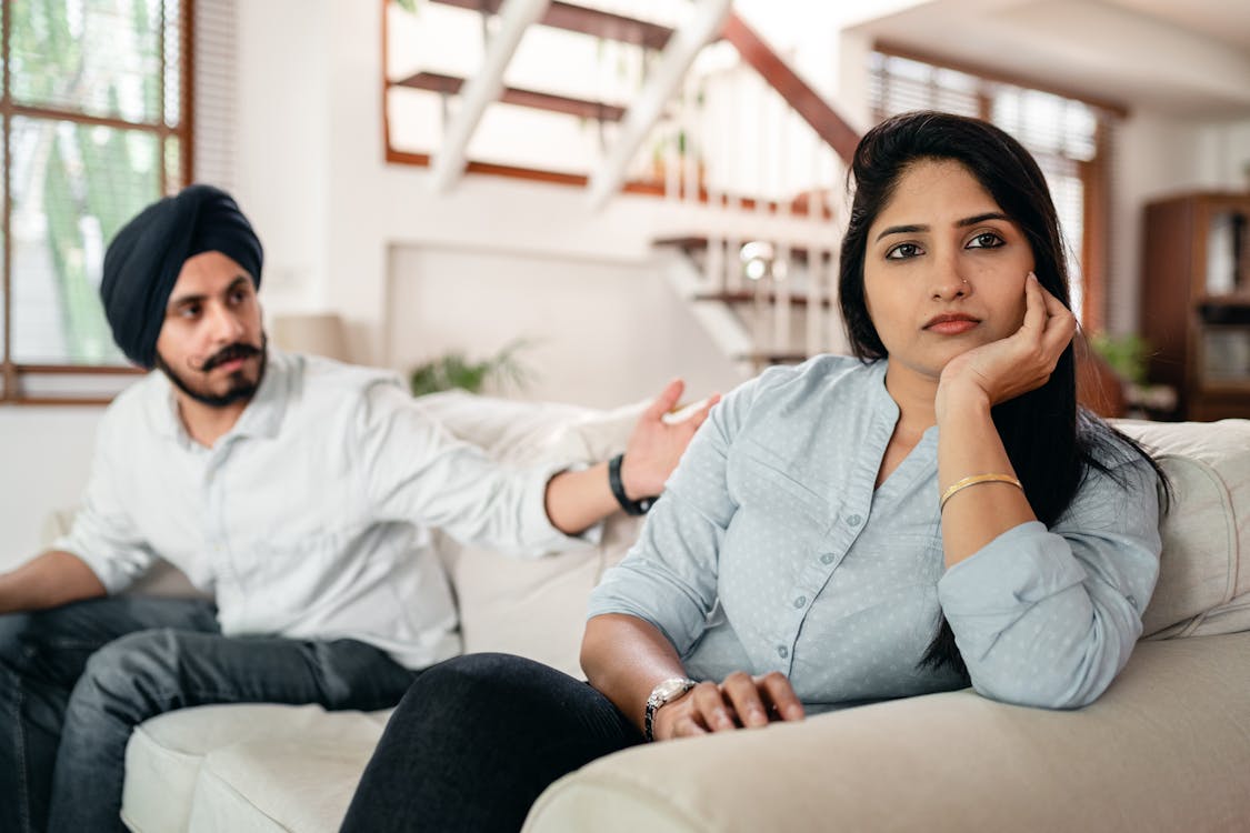 Free Young bearded Sikh man in casual wear and turban arguing with sad wife sitting on sofa Stock Photo