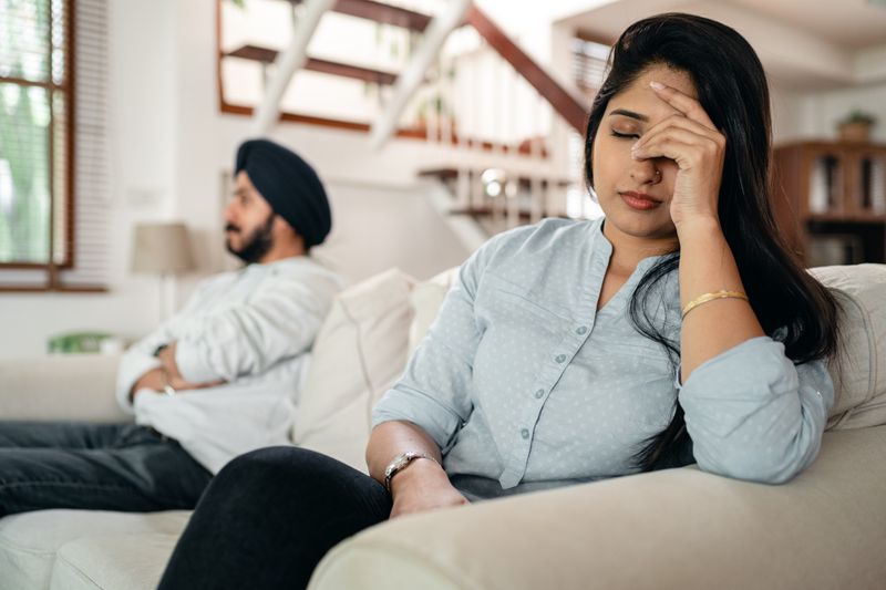 Tired frustrated young Indian woman in casual clothes having headache while sitting on sofa after arguing with husband