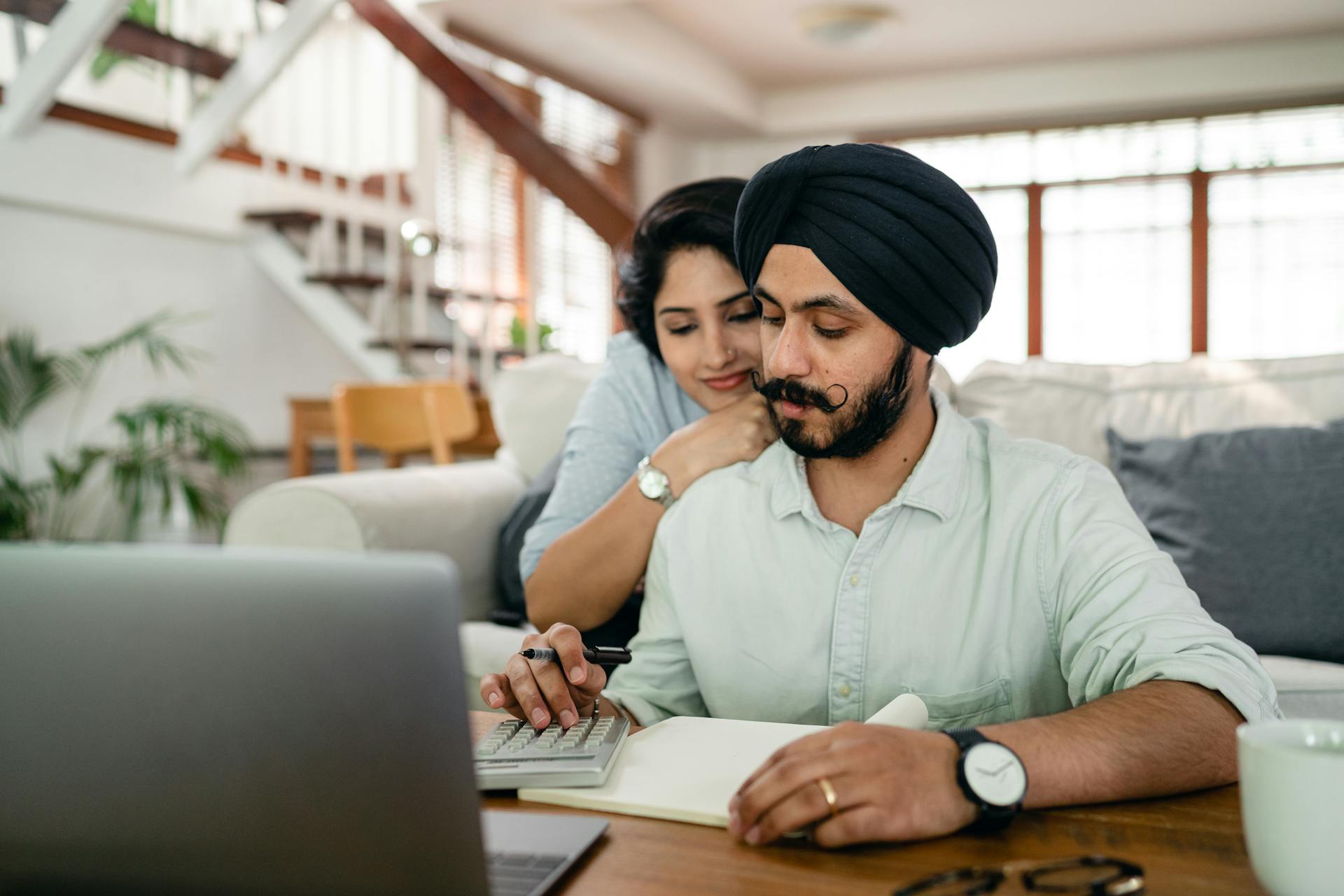 Young smiling woman hugging concentrated husband from behind while counting bills on calculator in modern apartment