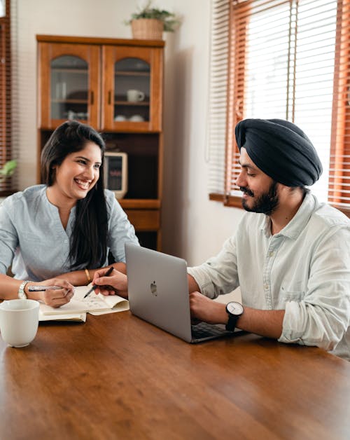 Smiling young Indian man in shirt and turban using laptop at wooden table and helping cheerful female friend while working remotely at home