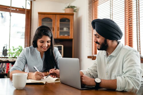 Smiling ethnic couple sitting at table and browsing laptop while writing notes in notepad and discussing business project together at home