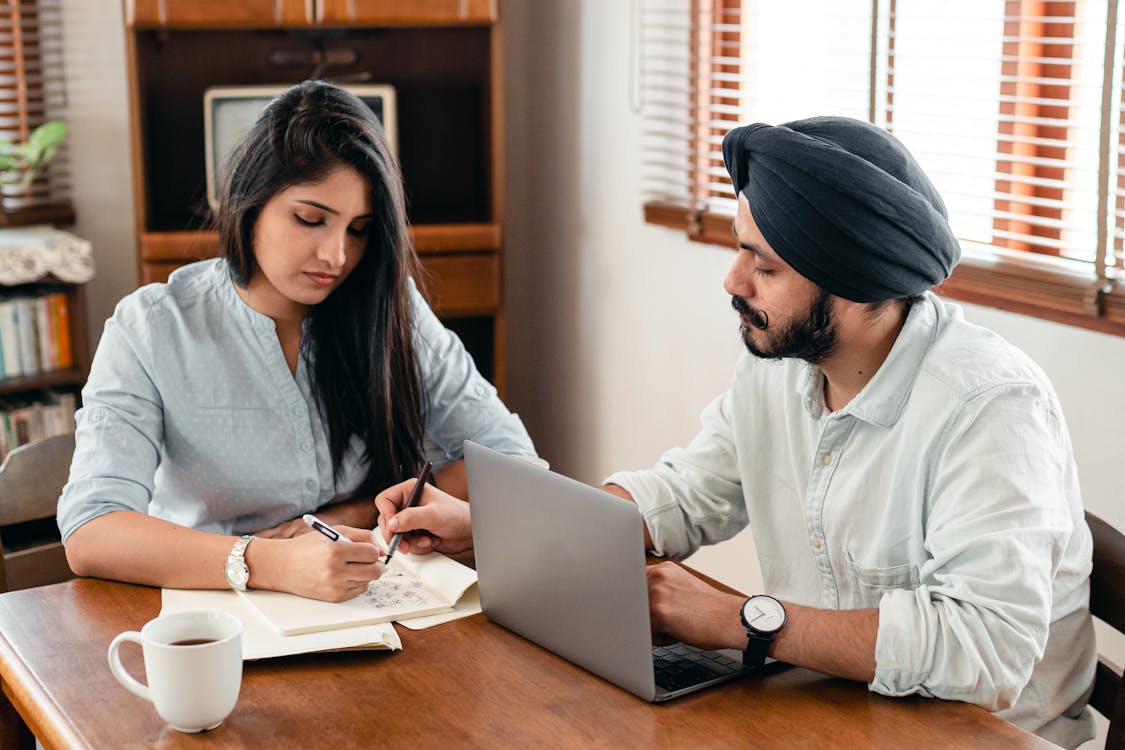 Free Indian man and woman sitting at table and working on notebook with bills and laptop while discussing business at home Stock Photo