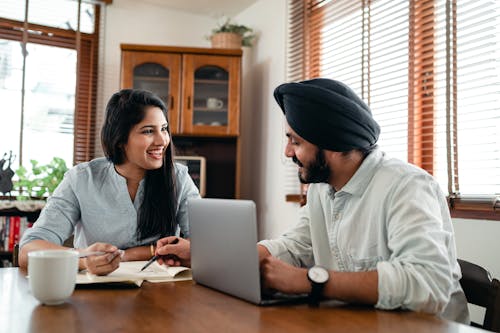 Free Laughing Indian woman with husband sitting at table and discussing project while working together with laptop and notebook at home Stock Photo