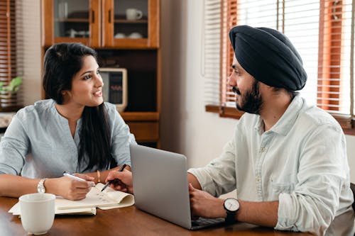 Indian male in turban and wristwatch typing on netbook while interacting with happy female coworker with notebook and pen sitting at table in office and looking at each other