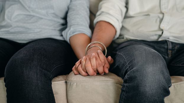一名穿着牛仔裤和衬衫、戴着腕带的匿名男子牵着女性的手，一起躺在公寓舒适的沙发上