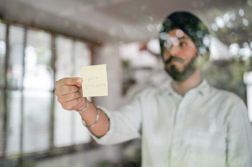 Indian guy in turban and shirt with curled mustache sticking paper with INDIA STAY HOME inscription while standing behind glass wall during COVID 19 pandemic