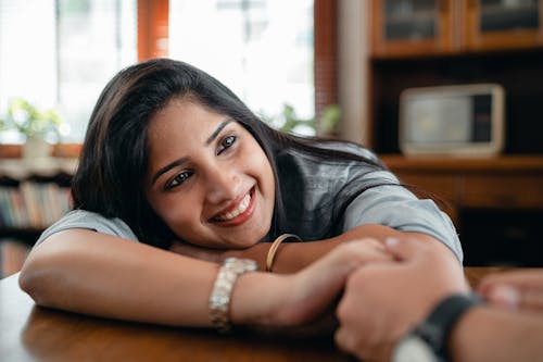 Cheerful Indian female in casual wear and wristband holding hand of crop faceless male beloved in wristwatch while sitting at table in living room in flat