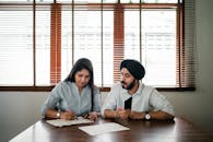 Indian woman with papers and pen writing on paper poster with ethnic Indian man in turban sitting nearby at wooden table