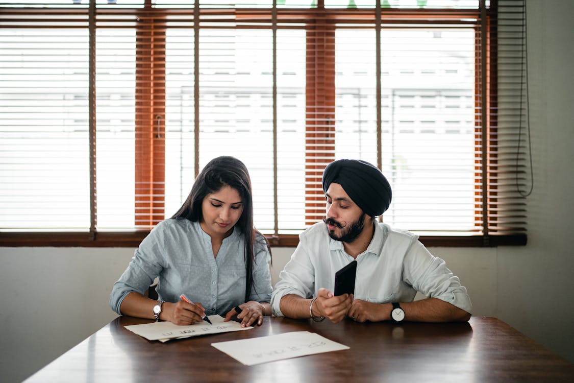 Free Indian woman with papers and pen writing on paper poster with ethnic Indian man in turban sitting nearby at wooden table Stock Photo