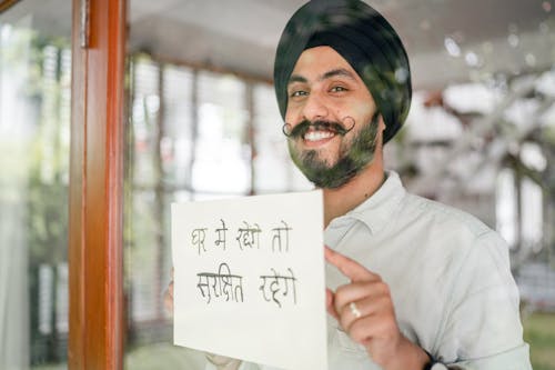 Free Positive mature modern Sikh male in turban with curly mustache leaning against glass wall with sign in indian on paper sheet and looking at camera Stock Photo
