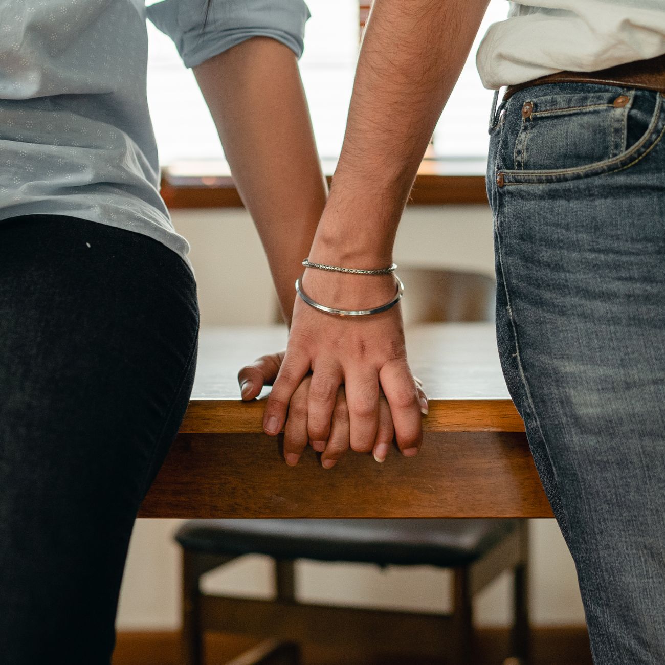 Crop anonymous young romantic couple in shirts and jeans holding hands while leaning on wooden table