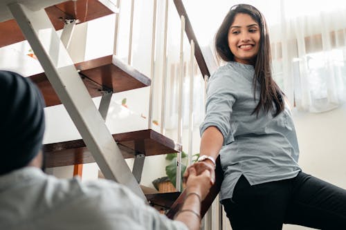 Free Smiling young ethnic lady holding hand of partner on stairs at home Stock Photo