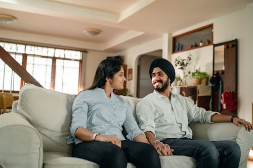 Happy smiling young Indian couple in similar outfits holding hands and looking at each other while resting on sofa in cozy living room