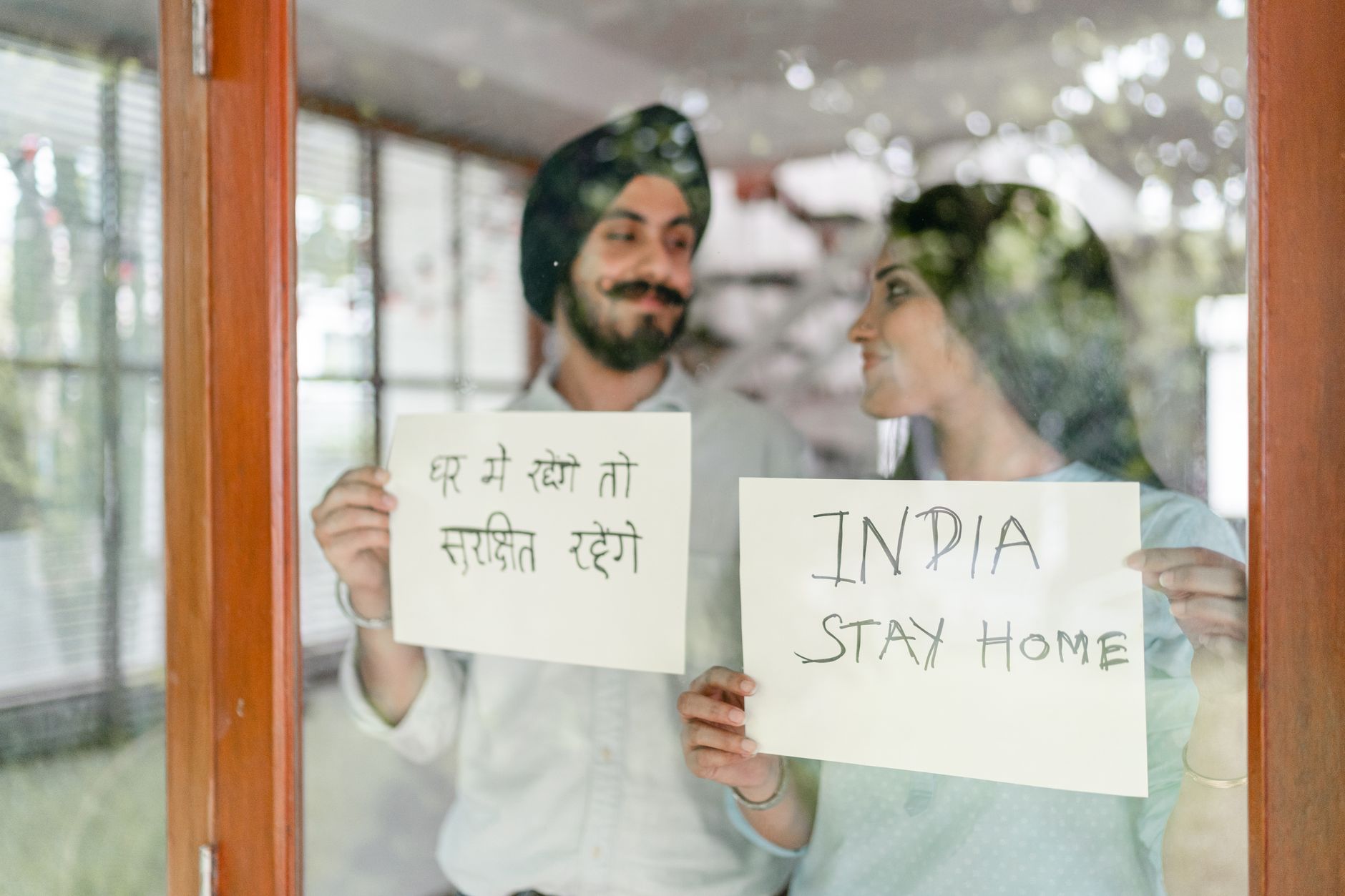 Positive young Indian couple in casual clothes standing against window and demonstrating sheets of paper with India stay home inscription during coronavirus