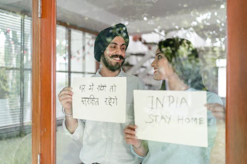 Through glass of young Indian couple standing against window and looking at each other while demonstrating sheets of paper with India stay home inscription in English and Hindi