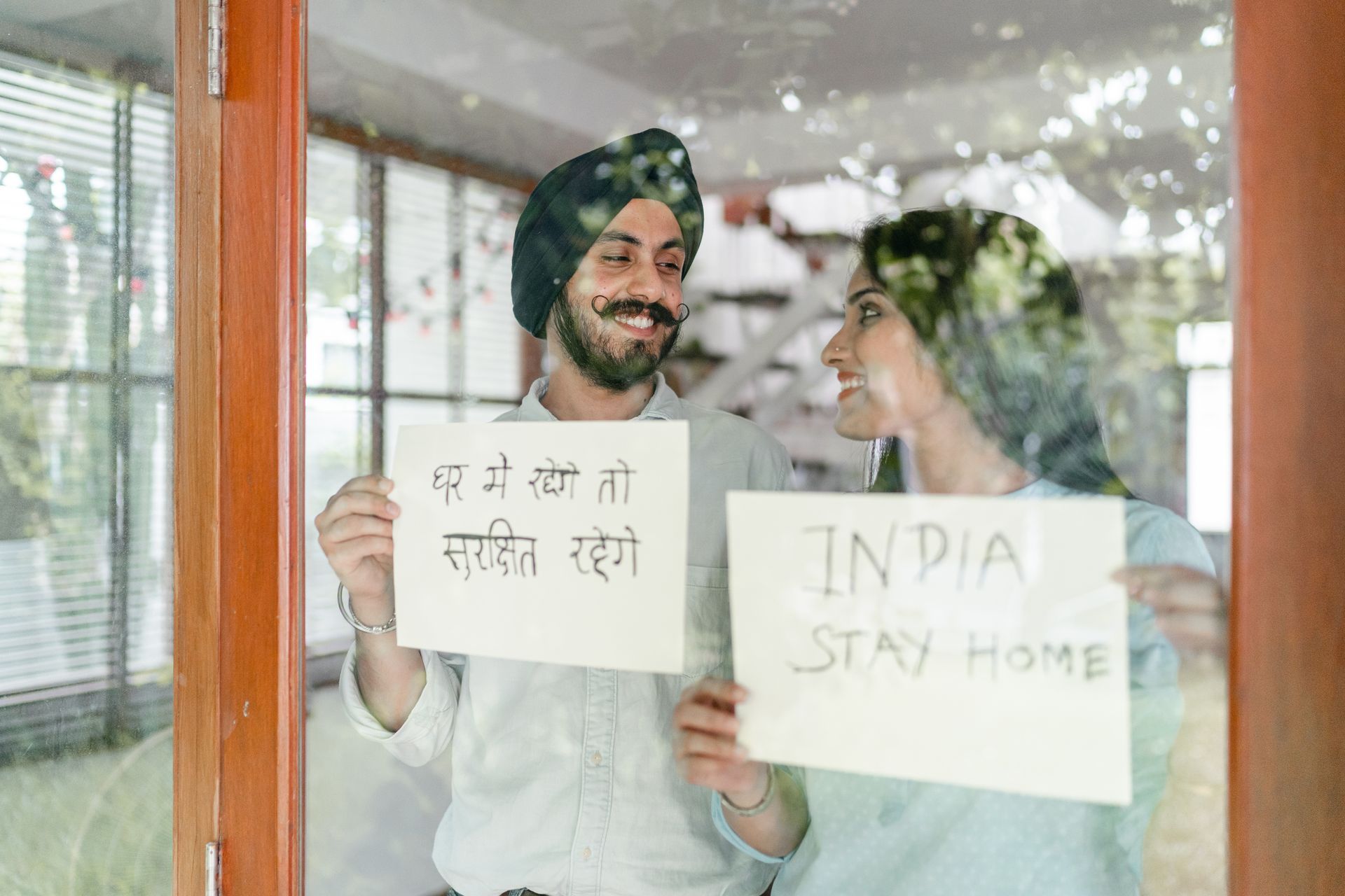 Through glass of young Indian couple standing against window and looking at each other while demonstrating sheets of paper with India stay home inscription in English and Hindi