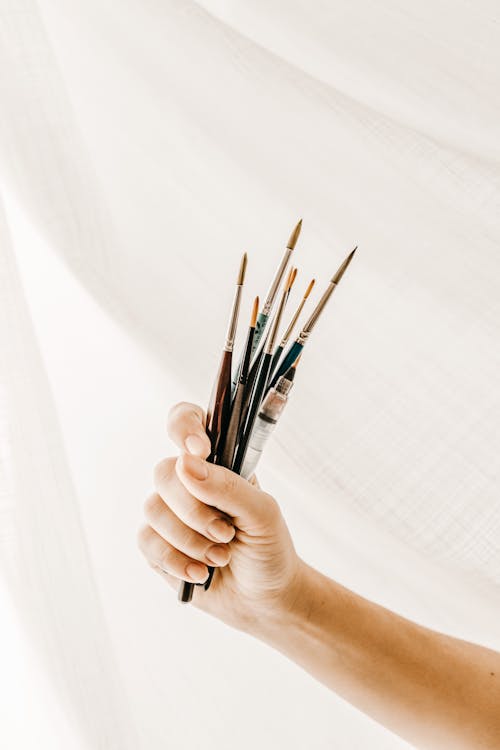 Free Unrecognizable artist showing set of paintbrushes against white drapery during work in studio Stock Photo