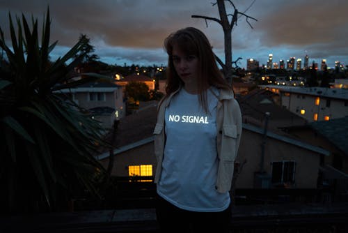 Calm female in casual white shirt standing on rooftop in residential area against cityscape and looking at camera seriously at night