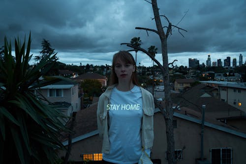 Young calm female in casual white shirt with illuminated stay home text standing on roof against night cityscape and looking at camera thoughtfully coronavirus concept