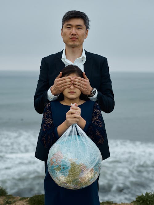 Asian male in formal suit standing against blurred sea covering eyes of emotionless girl in black dress with Earth globe in plastic bag environmental problems neglecting concept