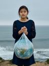 Unemotional Asian girl in black dress with Earth planet globe in plastic bag standing against blurred cloudy seashore environmental pollution problems concept