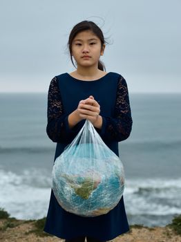 冷漠的亚洲女孩穿着黑色连衣裙，拿着塑料袋里的地球仪，反对关于模糊的多云海滨环境污染问题的概念