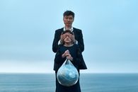 Asian male in formal wear standing near seashore and covering eyes of girl in black dress with Earth globe in plastic bag environment pollution concept