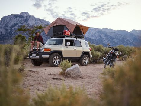 Outdoor camping scene with a man sitting on a car equipped with a rooftop tent, scenic mountain view.