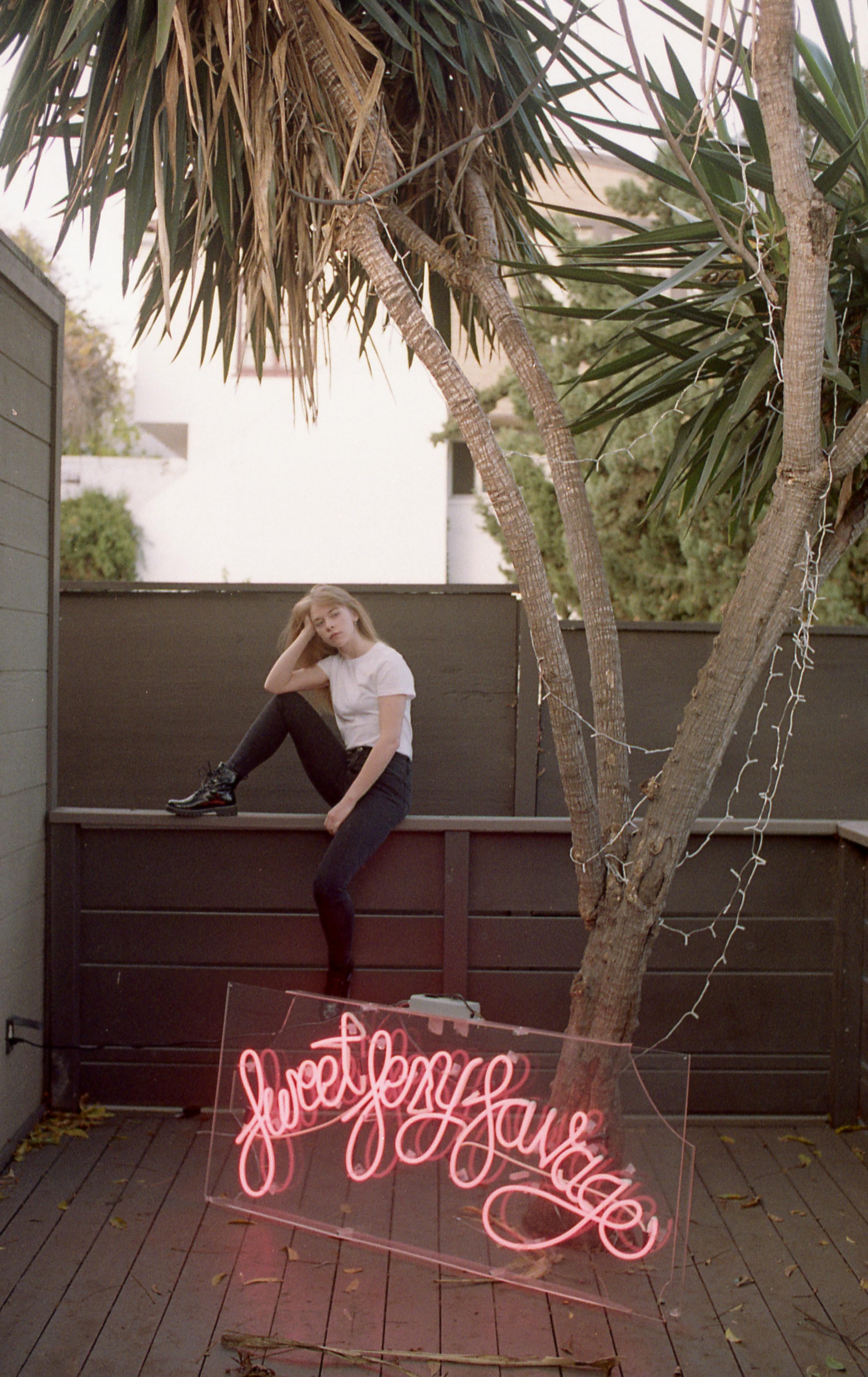 Slim young lady in trendy outfit sitting alone on wooden bench on porch near neon letters and trees