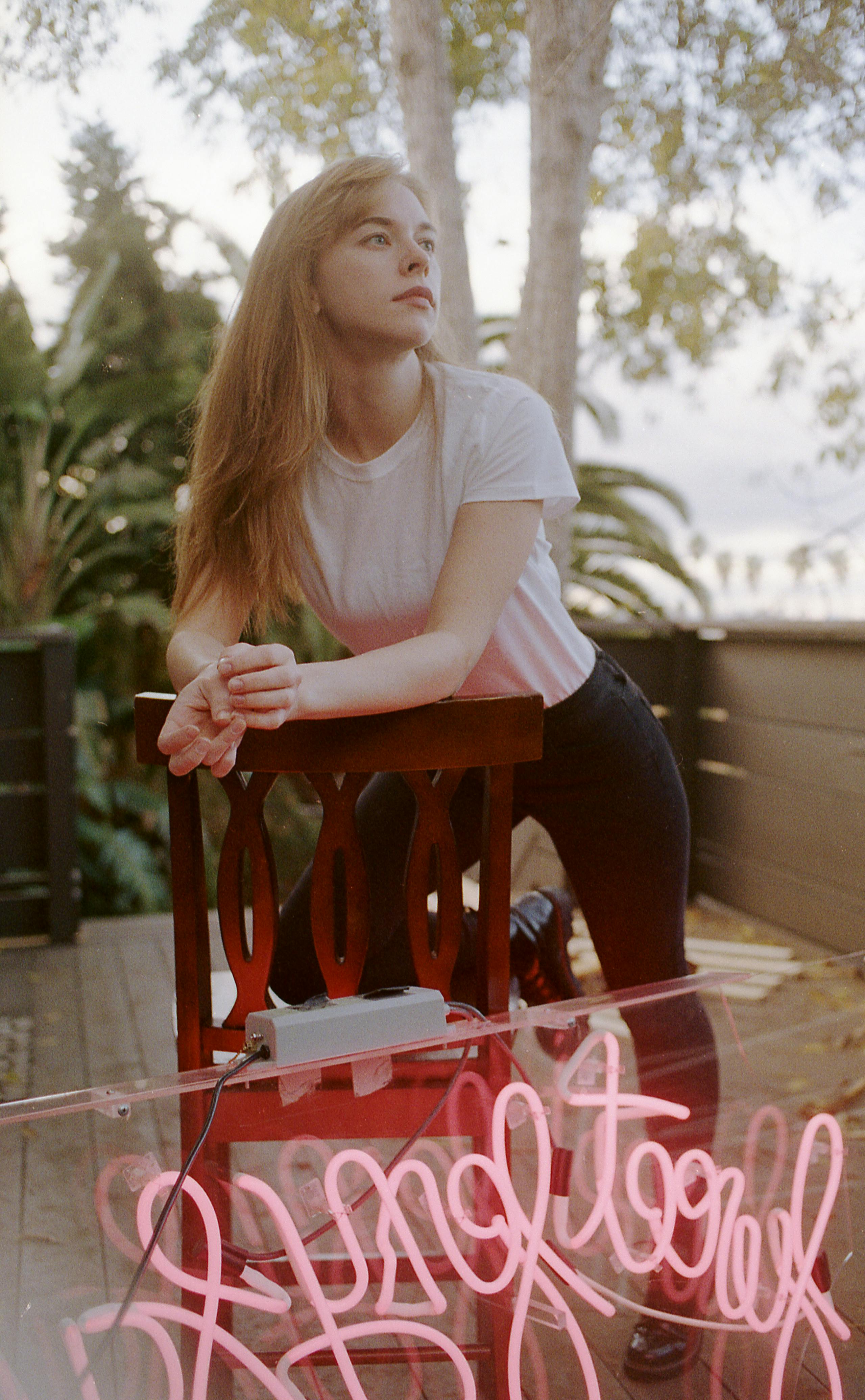 A woman in a white shirt leans on a chair outdoors, next to a pink neon sign.